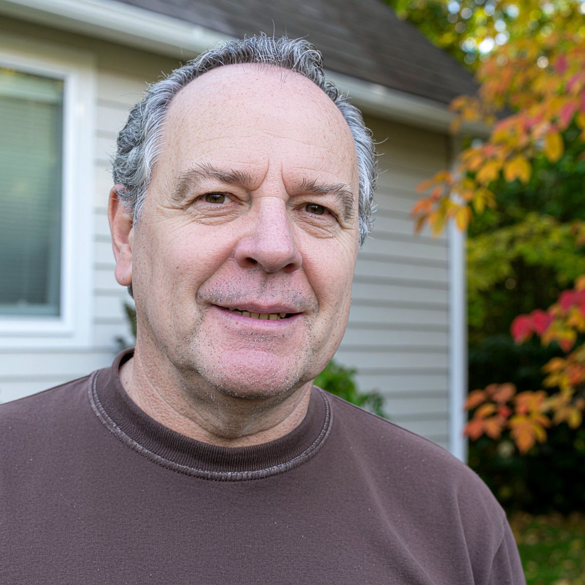 Gray-haired man wearing a brown sweatshirt standing outside of his home.