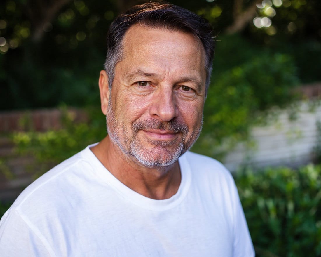 Man in white t-shirt outside in front of short brick wall and trees.