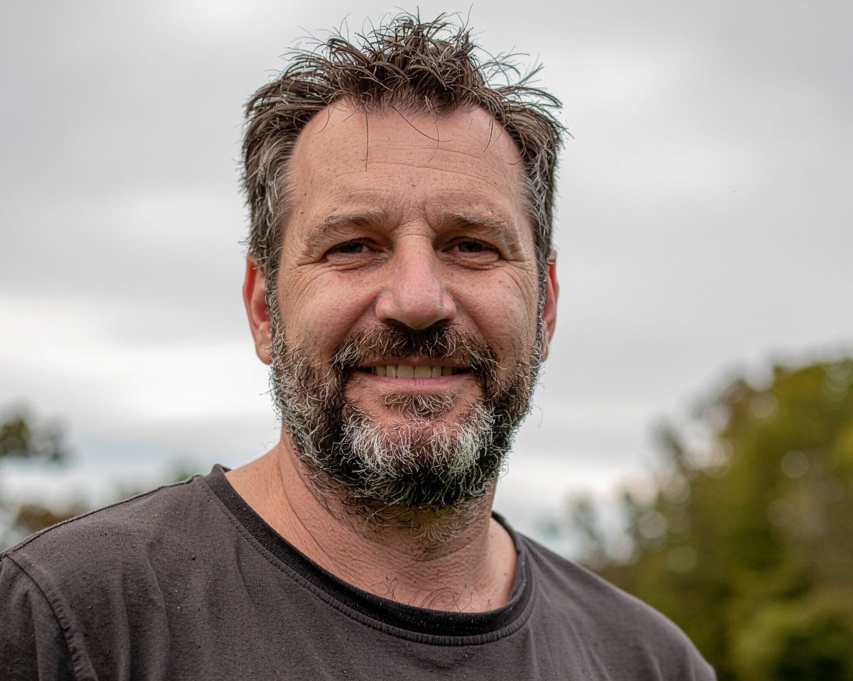 Middle aged man with a scruffy beard wearing a gray t-shirt standing outside with out of focus trees and sky in the background.