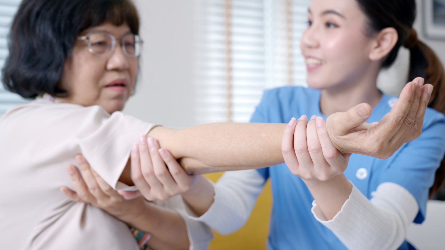 Young nurse in blue scrubs attending to middle aged woman with arm extended.