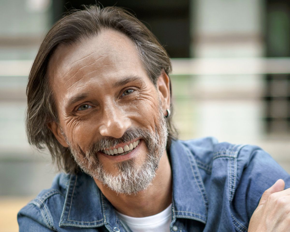 Smiling man wearing a white t-shirt and a bluejean short over it unbuttoned sitting in front of an out of focus building in the background.