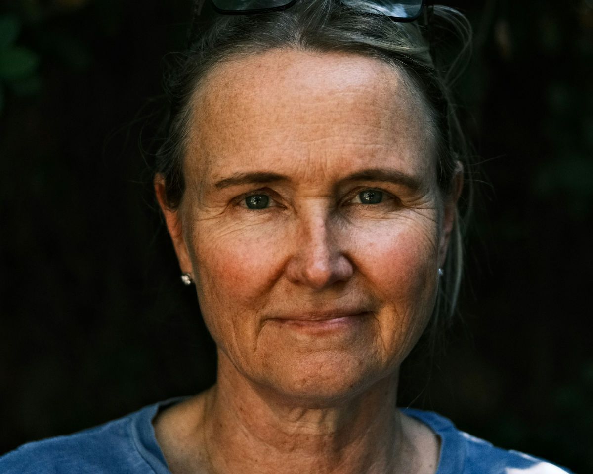 Woman wearing blue shirt and glasses pushed up in ther hair, standing outside in front of trees.
